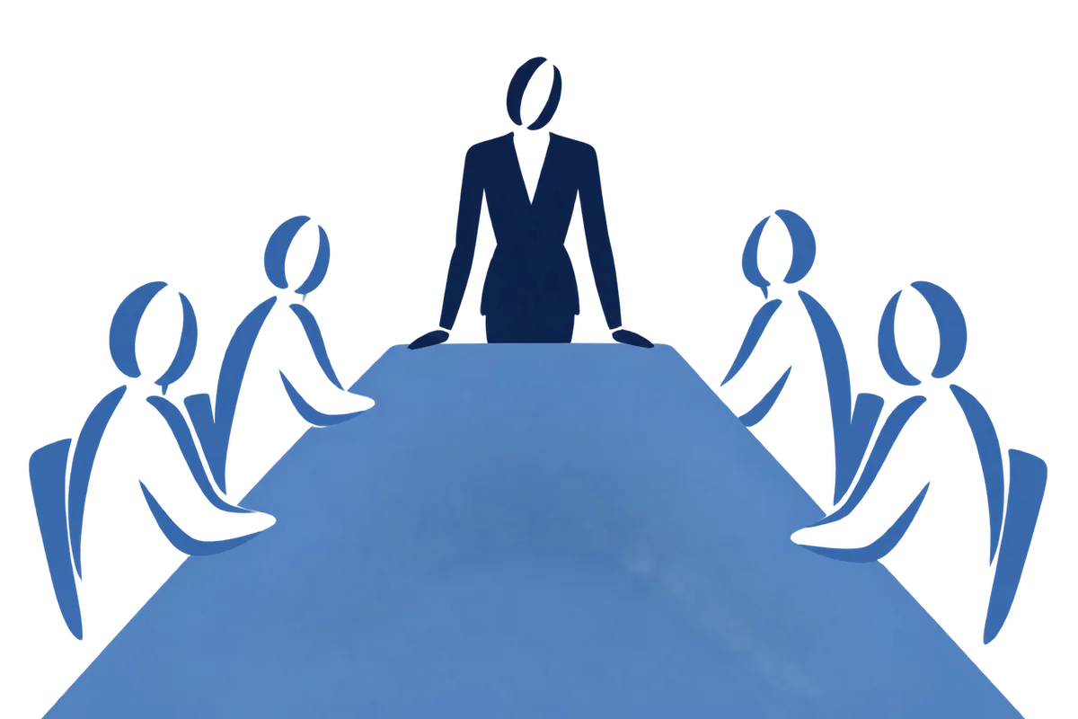A person standing at the head of a long table, others seated and turned towards them.